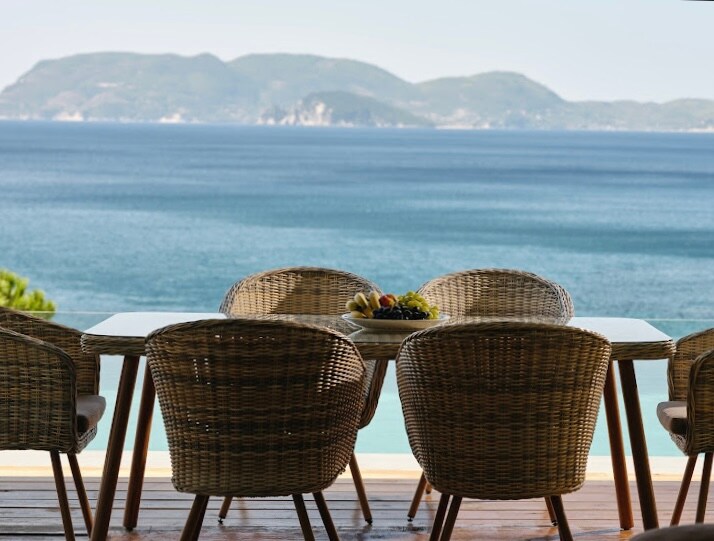 An outdoor dining area features a wooden table accompanied by four woven chairs. A fruit arrangement is placed at the center of the table, with expansive views of the sea and distant islands visible in the background.