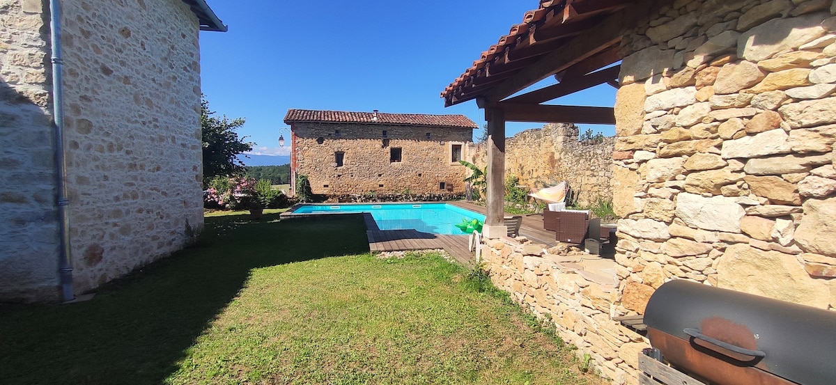 A spacious outdoor area features a well-maintained swimming pool, surrounded by a verdant lawn and rustic stone walls. Nearby, a covered patio provides seating and a barbecue grill, while a charming stone building is visible in the background.