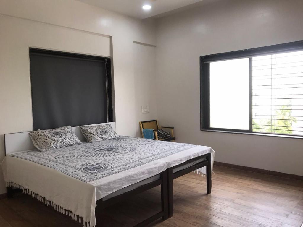 A bright sleeping area is centered by a spacious bed adorned with a patterned bedspread. Natural light enters through a large window, which is equipped with horizontal blinds. A chair is positioned beside the window, while the wooden floor adds warmth to the room.