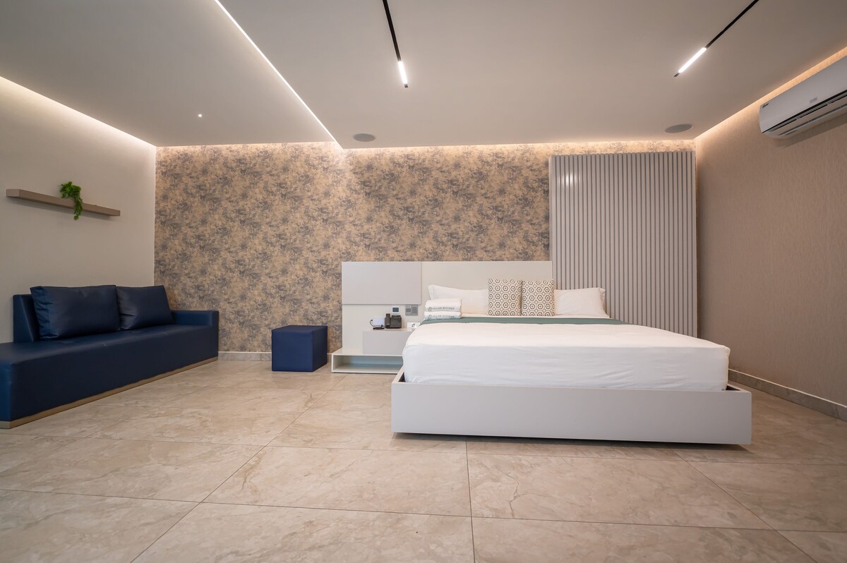 A spacious room features a large bed with a simple headboard, complemented by a sleek sofa and an accent table. Soft lighting highlights the modern design, and light-colored walls provide a serene backdrop. A cool tile floor adds to the contemporary feel.