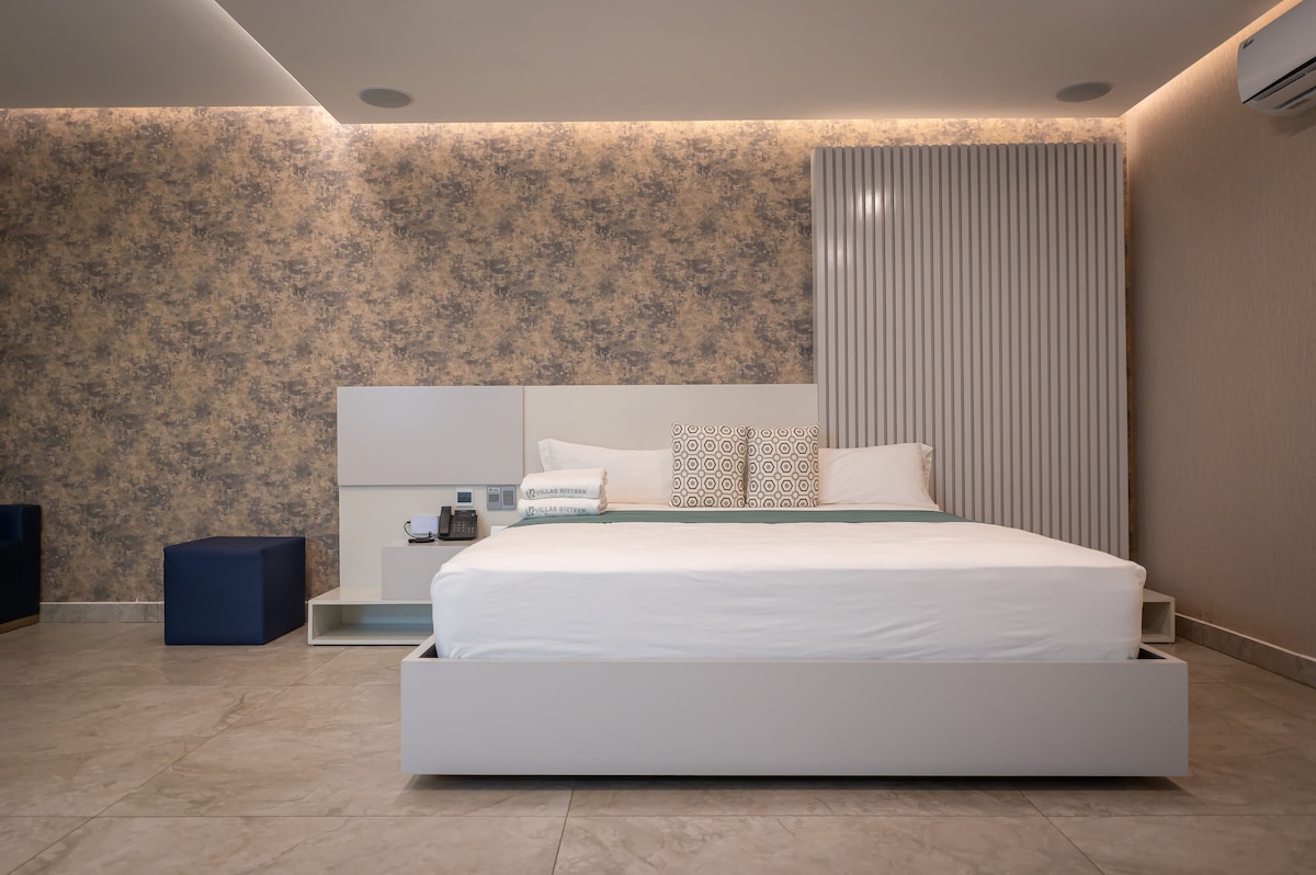 A spacious bedroom features a modern design with a large bed centered against a textured wall. Decorative pillows are arranged at the head of the bed, complemented by minimalist side tables. Soft lighting creates a serene ambiance, while tile flooring adds a touch of elegance.