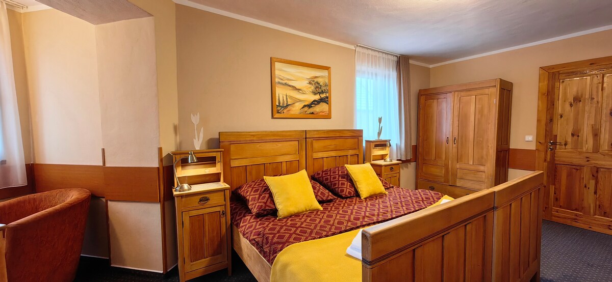 The bedroom features a wooden double bed with decorative pillows, paired with matching bedside tables and lamps. A wardrobe occupies one corner, and a comfortable chair is positioned nearby. Natural light enters through a window adorned with sheer curtains, accentuating the room's warm tones.