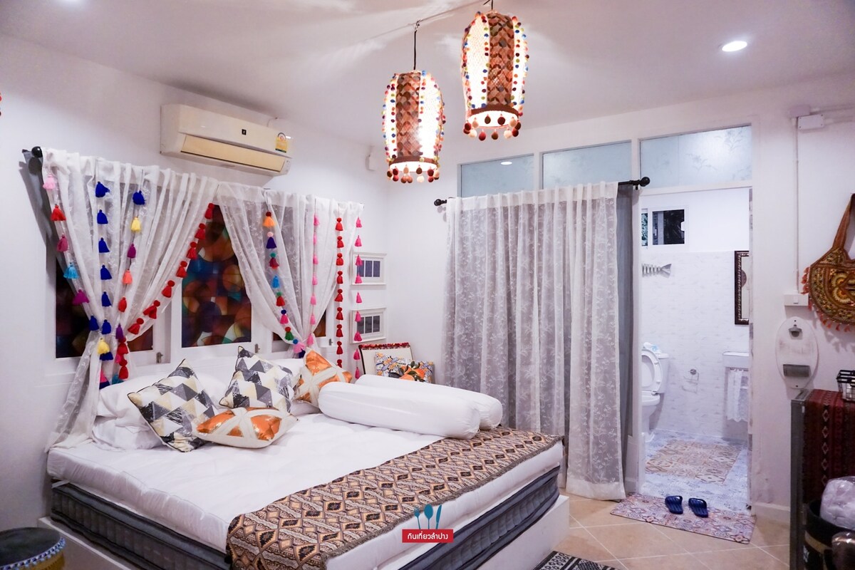 A comfortable bedroom is designed with a blend of colorful textiles and decorative lanterns. A spacious bed is adorned with patterned pillows and a white blanket. Sheer curtains frame the window, and a private bathroom doorway is visible in the background.