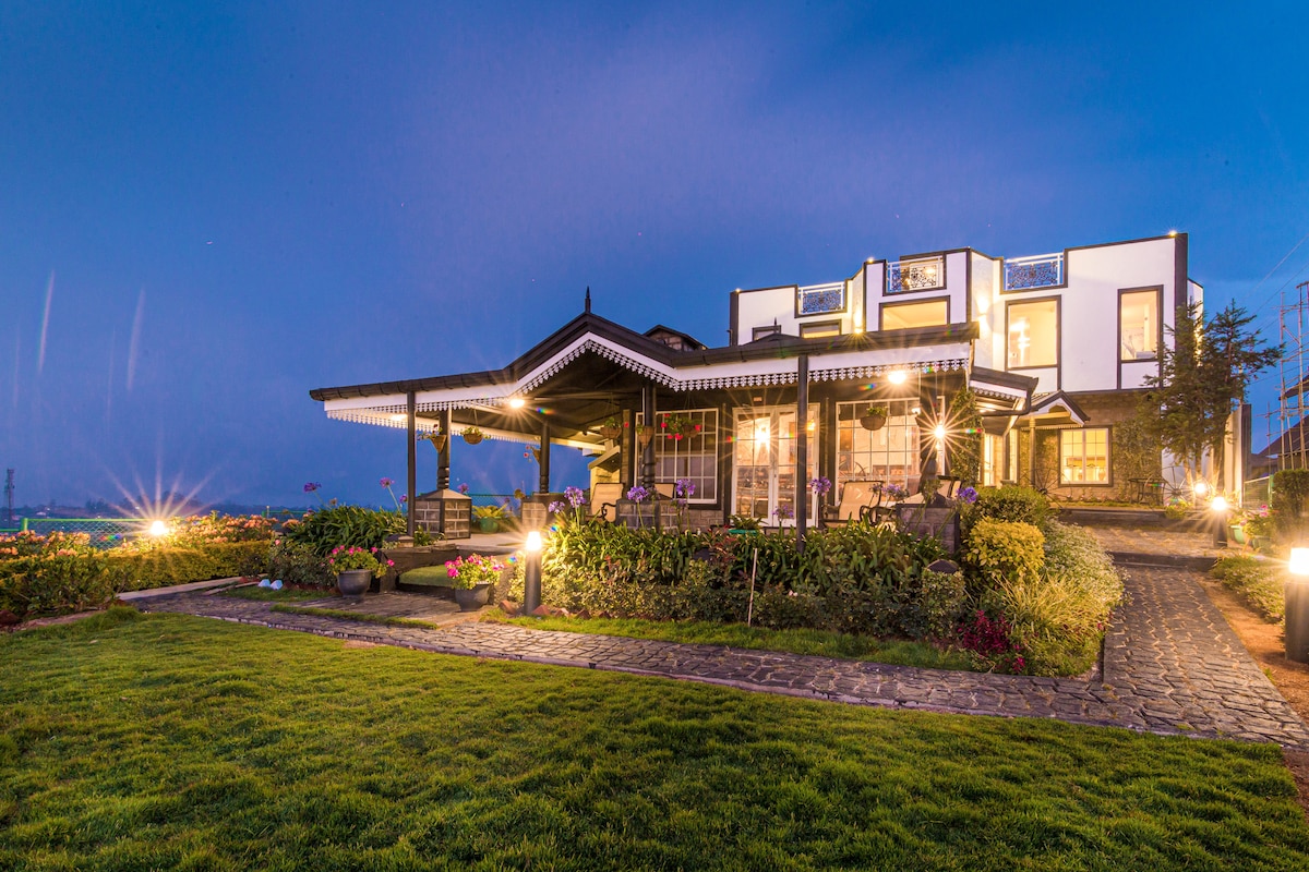 The bungalow's exterior is illuminated at dusk, highlighting the elegant architecture and vibrant gardens. A spacious patio features a covered seating area, surrounded by neatly trimmed grass and colorful flowers, creating a tranquil space for relaxation. Surrounding views of nature are visible in the background.