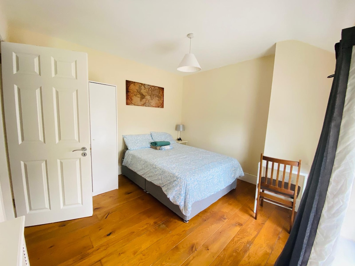 A well-lit double room features a spacious bed covered with a blue-patterned duvet. Wooden flooring enhances the warmth of the space. A small bedside lamp and a wall art piece of a map add to the decor. A wooden chair is positioned near the window, with a wardrobe visible.