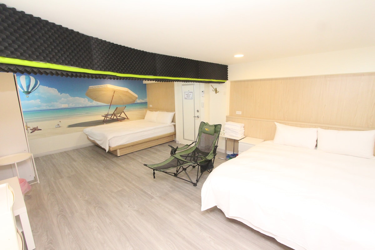 A spacious room features two large beds with white linens and a comfortable folding chair. A mural depicting a serene beach scene adorns the wall, while light wood accents complement the neutral flooring. Natural light filters in, enhancing the overall brightness.