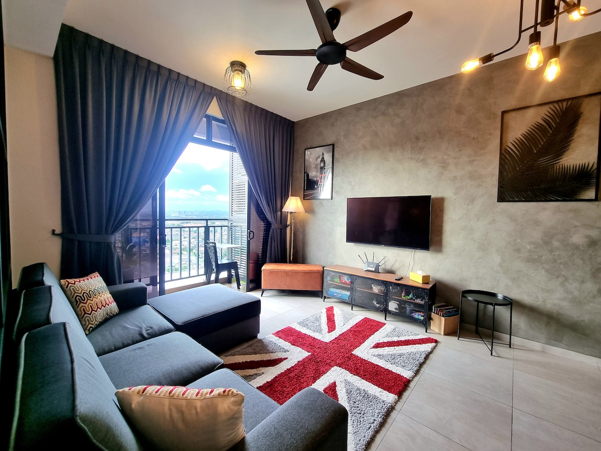 A modern living area features a gray sofa adorned with colorful cushions and a plush rug depicting the Union Jack. Large windows allow abundant natural light, complemented by dark curtains. A wall-mounted television is positioned on a media console, with an accent chair visible in the corner.