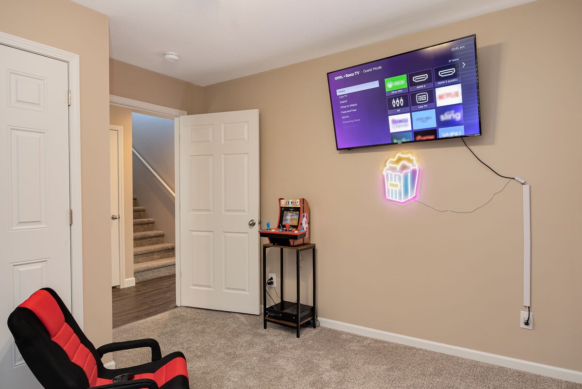 A cozy entertainment area is showcased with a comfortable chair facing a wall-mounted television. A small side table holds a variety of gaming consoles. Neutral wall tones provide a calm backdrop, and soft carpet flooring enhances the comfort of the space.