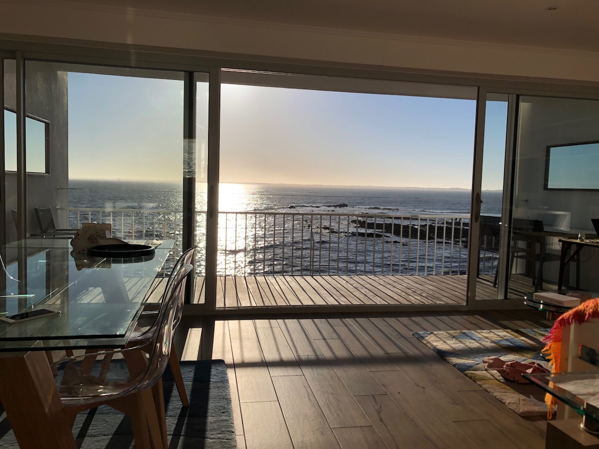 A spacious living area is captured, featuring large glass sliding doors that offer unobstructed views of the ocean. The warm glow of sunset reflects on the water, enhancing the serene ambiance. A glass dining table and a brightly colored throw blanket are visible in the foreground.