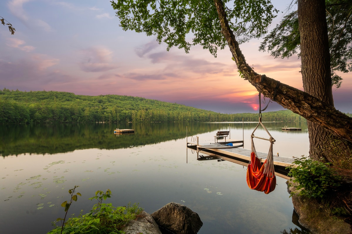 A serene lakeside view captures a tranquil setting at sunset, with a gentle reflection of trees and sky on the water. A hammock hangs from a sturdy tree, adding to the peaceful ambience. A wooden dock extends into the calm water, where kayaks and boats are anchored.