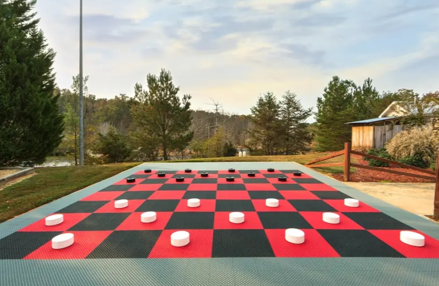 An outdoor checkers board is displayed on a large rubber mat, featuring alternating red and black squares. The game pieces are arranged on the board, surrounded by trees and a peaceful landscape in the background, indicating a recreational area for guests to enjoy.