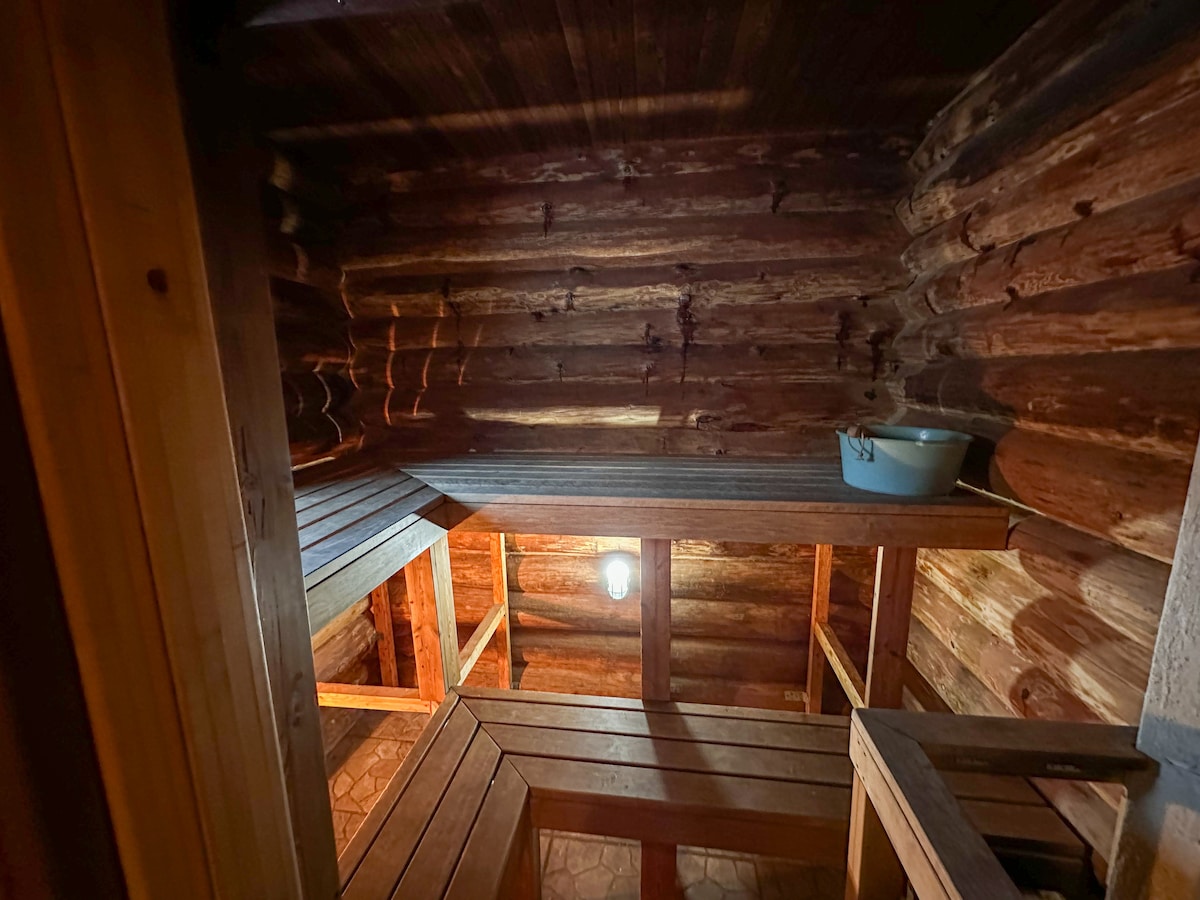 The interior of the sauna features natural wooden logs, creating a warm, rustic atmosphere. Wooden benches are arranged for seating, complemented by soft lighting from a ceiling fixture. A small basin sits on one of the shelves, enhancing the functional space.