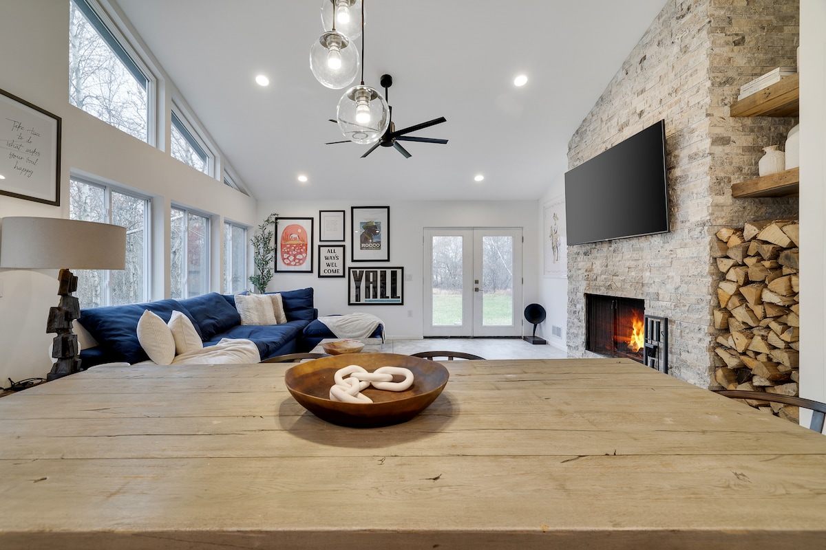 An open-concept living area showcases a rustic dining table, complemented by a textured bowl at the center. A modern fireplace rests against a stone wall, while large windows allow natural light to fill the space, highlighting a cozy lounge area with blue sofas.