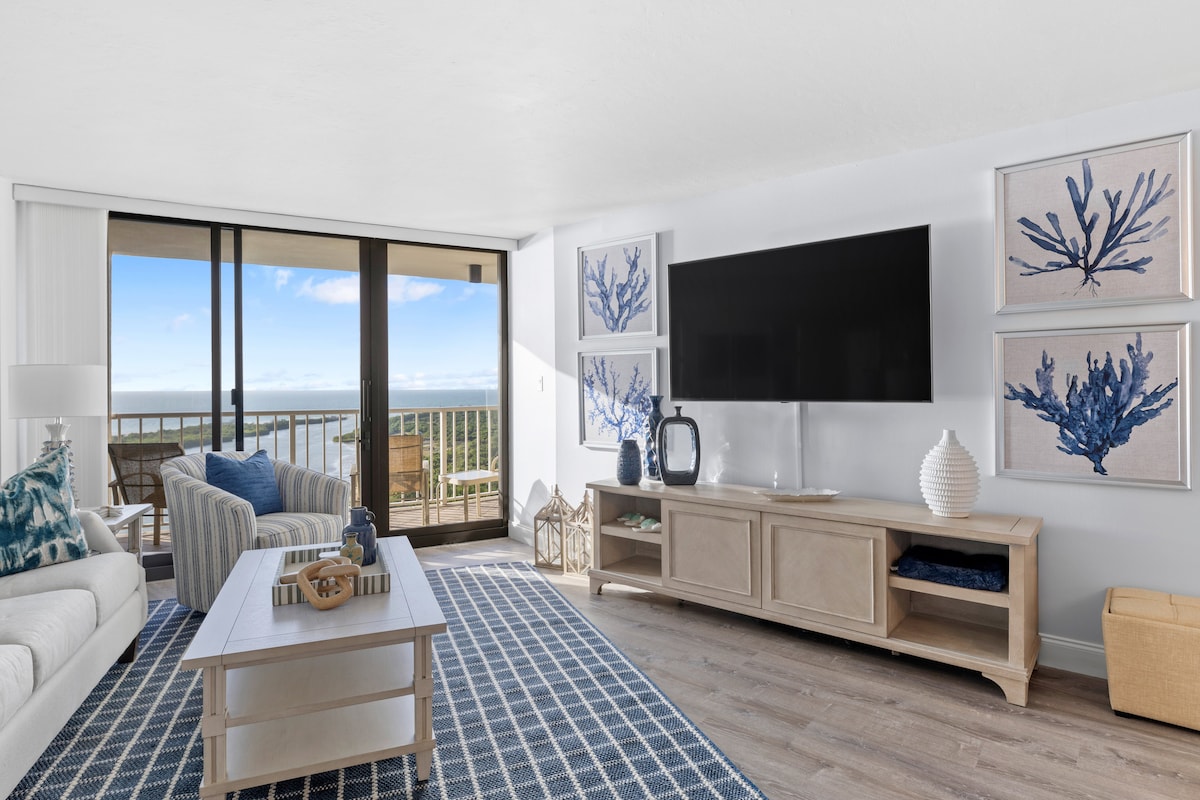 A bright living area features a comfortable white sofa and a patterned blue area rug. Large glass doors frame the expansive view of the Gulf of Mexico. Coastal-themed artwork adorns the walls, and a flat-screen TV is mounted above a light wooden media console.