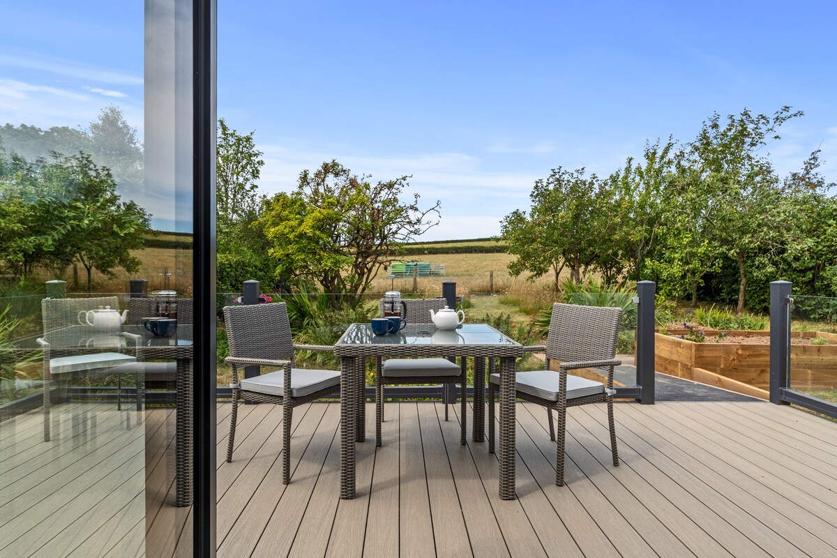 A private terrace is featured with a glass balustrade and a table set for four. Surrounding greenery adds natural beauty, with farmland visible in the background under a clear blue sky.