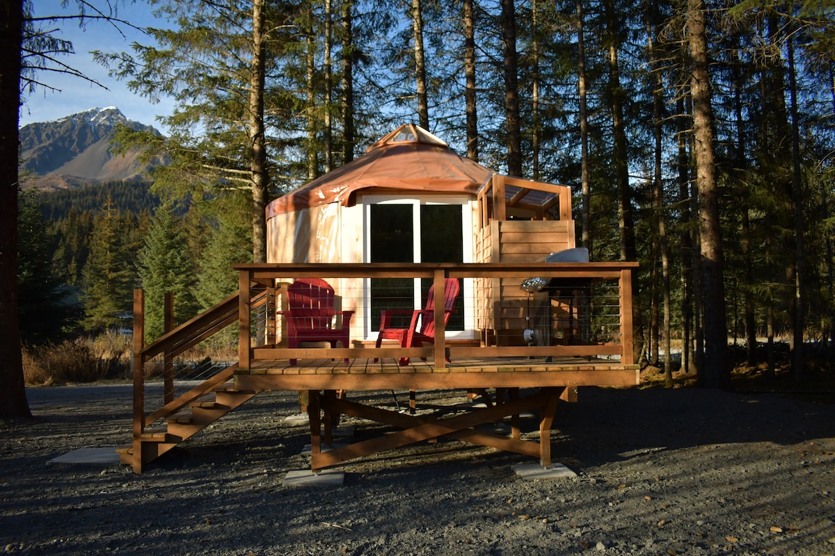 A wooden deck leads to the 16ft yurt, situated among spruce trees. Red chairs are positioned on the porch, offering a view of the surrounding natural landscape. The exterior features soft colors, blending harmoniously with the environment, and a wooden staircase provides easy access.