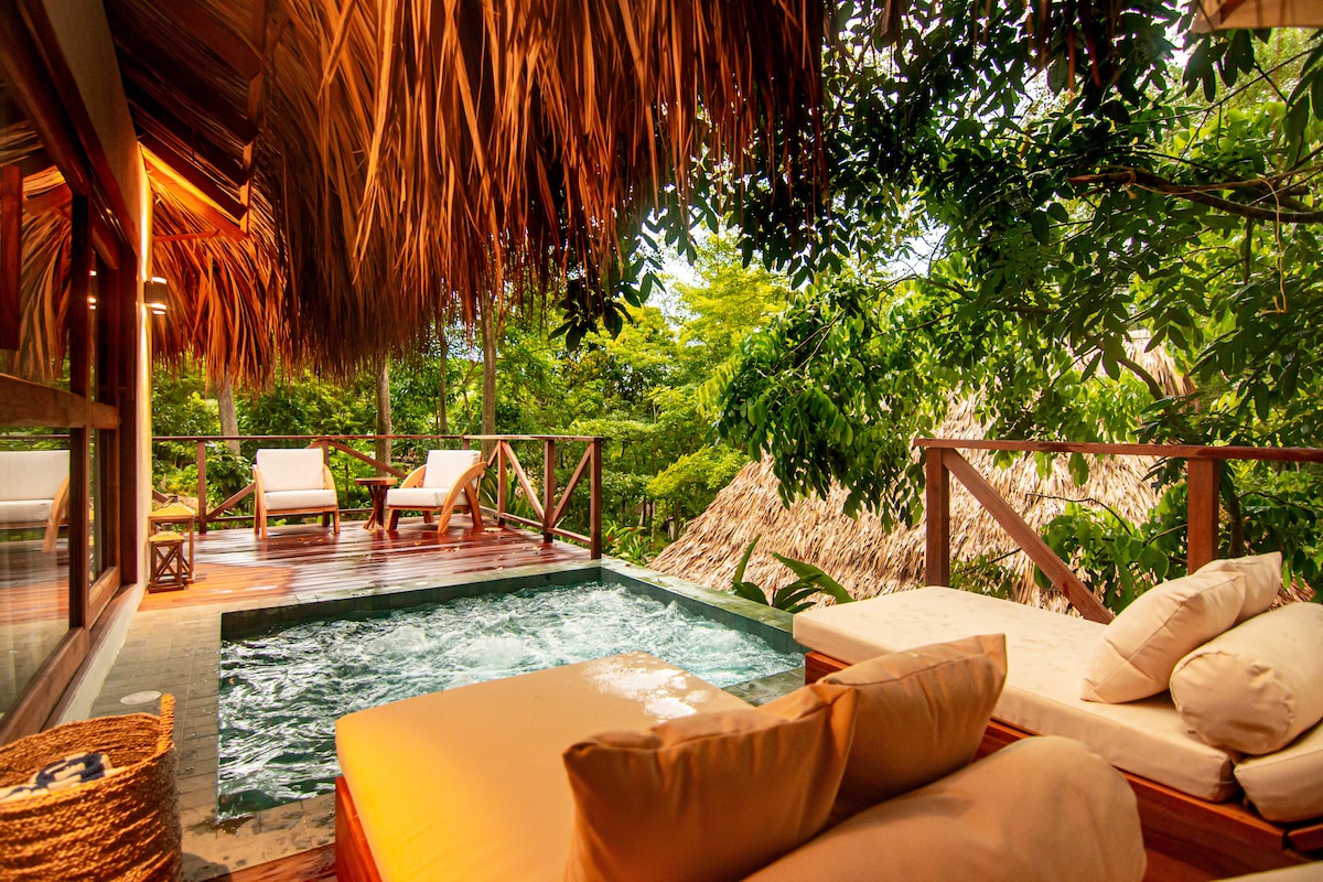 A private outdoor jacuzzi is surrounded by lush greenery, with a thatched roof providing shade. Two sun loungers are positioned nearby on the wooden deck, inviting relaxation amid the serene natural setting.