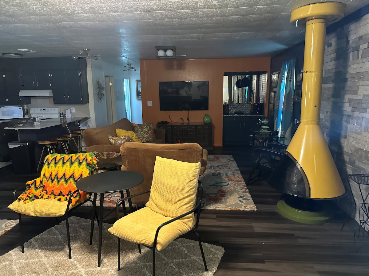 A vintage living room showcases a mix of retro furniture including a brown seating area and colorful armchairs. A yellow freestanding fireplace provides a focal point against a stone wall. A flat-screen TV is mounted, and a patterned area rug adds texture to the warm space.