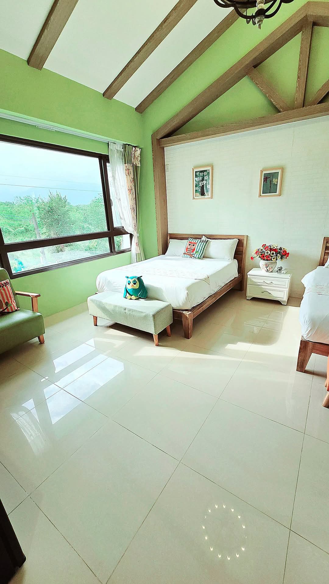 A bright, airy room features two beds, one with a soft footstool. Large windows allow natural light to fill the space, showcasing scenic views of greenery outside. Neutral wall tones are complemented by wooden beams and colorful accents, creating a warm, inviting atmosphere.