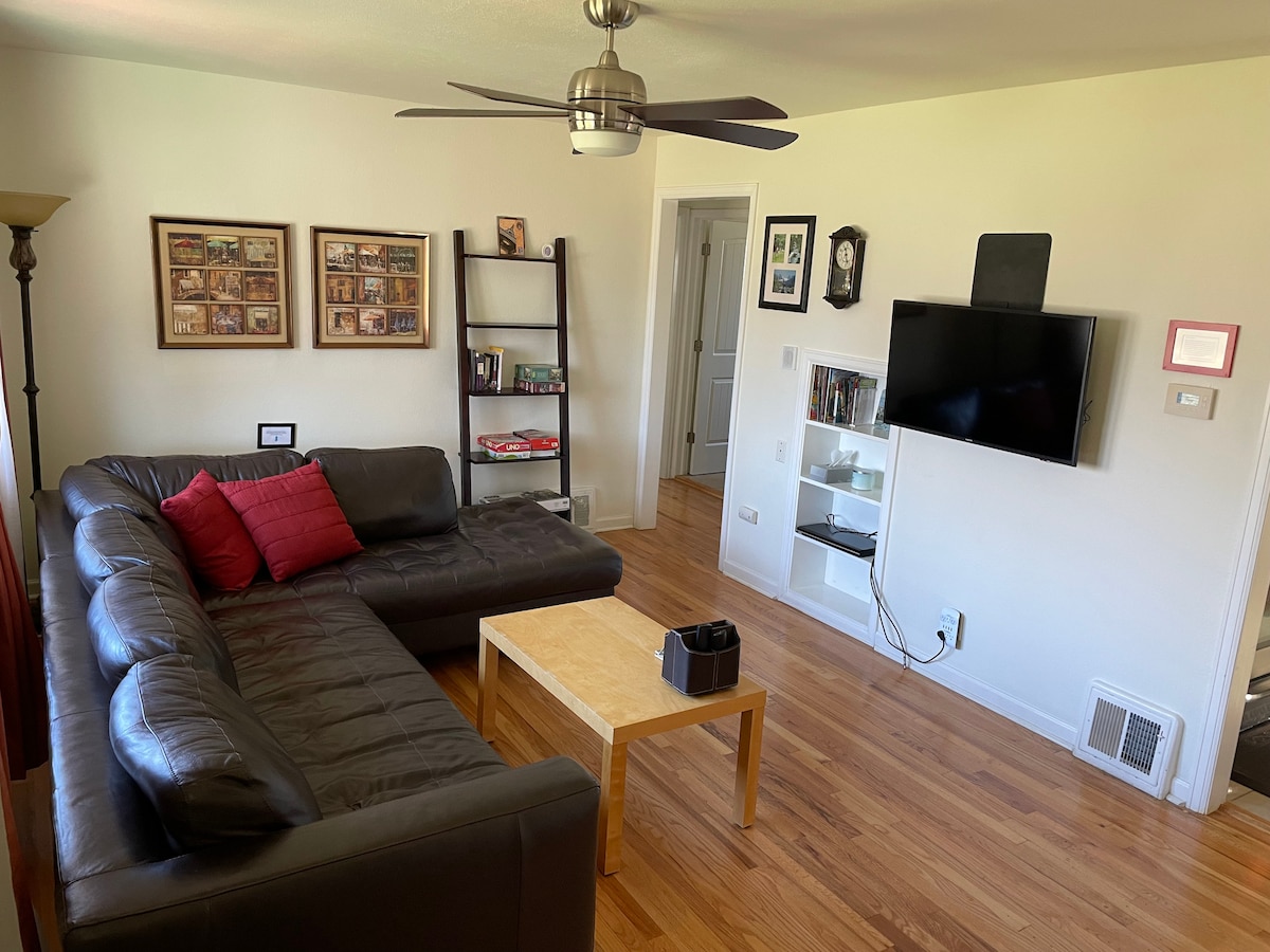 A comfortable living room features a dark leather sectional sofa complemented by red accent pillows. A light wooden coffee table is positioned in front, and a mounted television is visible on the wall. A bookshelf displays various items, enhancing the room's cozy ambiance.