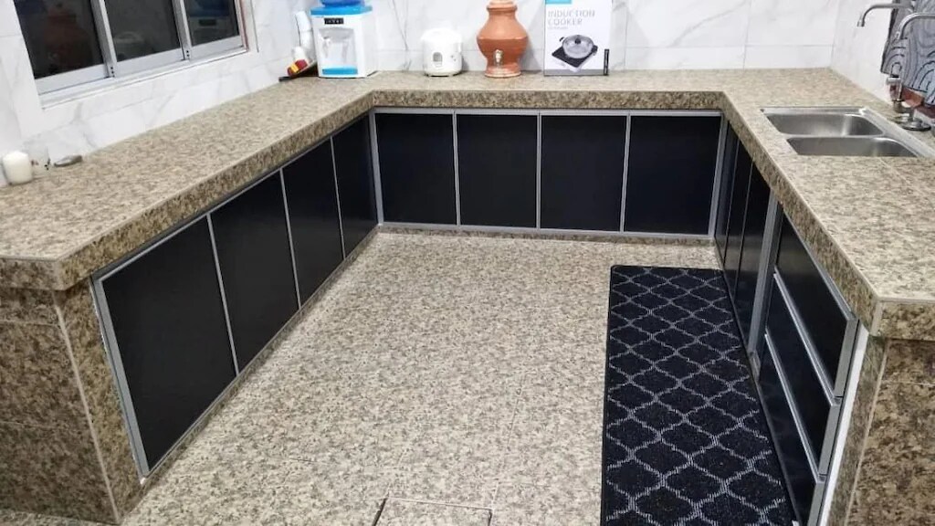 A modern kitchen space is presented with a large granite countertop and sleek black cabinetry. The flooring features a patterned rug for comfort, and natural light enters through a nearby window, enhancing the functional layout, which includes a double sink.