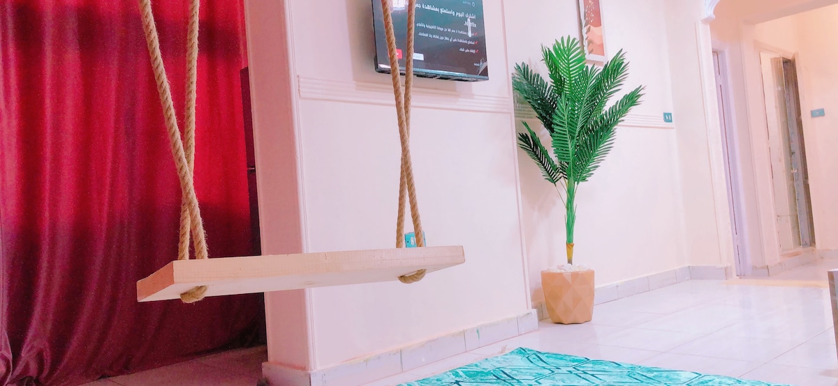 A cozy living area is highlighted by a wooden swing hanging from the ceiling. A decorative plant in a textured pot adds a touch of greenery, while a vibrant area rug complements the soft color tones of the space. A TV is mounted on the wall near a rich red curtain.