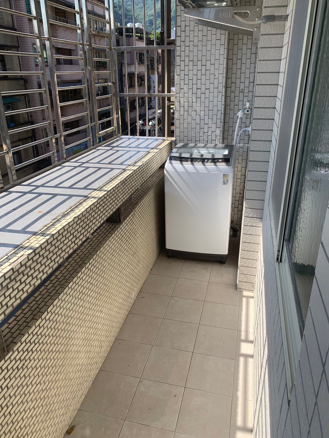 A compact outdoor balcony is showcased, featuring tiled flooring and a minimalist design. A white washing machine is positioned against a textured tiled wall, while a railing offers an unobstructed view of the surroundings. Natural light brightens the space through a nearby window.
