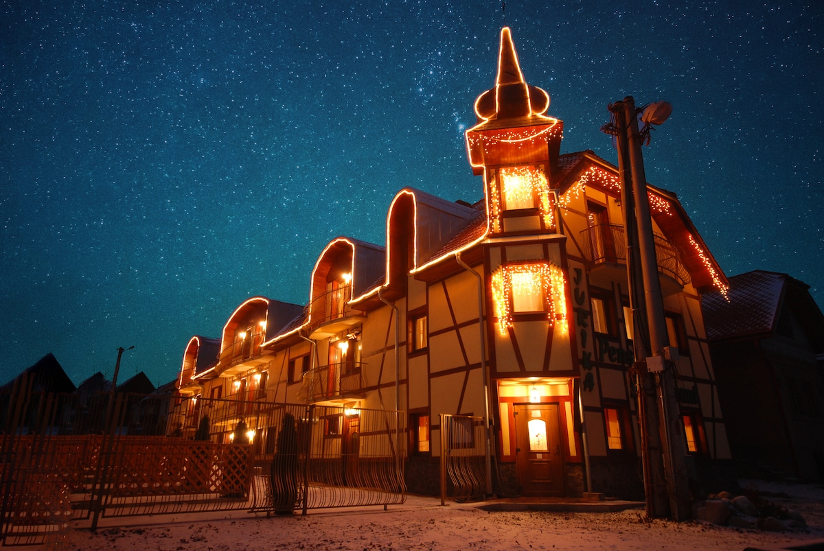 A three-story building is illuminated with warm lights against a starlit sky. The structure features a distinctive architectural style, with a decorative turret and multiple gabled roofs. The surrounding area is clear, highlighting the building's unique design.