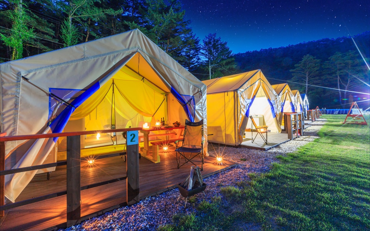 Several brightly lit glamping tents are arranged along a grassy area, with wooden decks and chairs positioned outside. Warm light glows from inside the tents, enhancing a cozy ambiance under a starlit sky. Trees are visible in the background, adding to the serene environment.