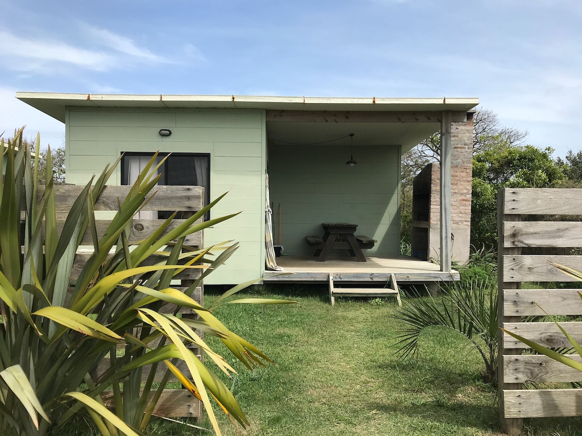 A charming cabin features a covered porch with a picnic table and a light fixture overhead. The structure is surrounded by lush greenery, and wooden fencing adds a rustic touch. The exterior is painted in a soft green, blending harmoniously with the natural surroundings.