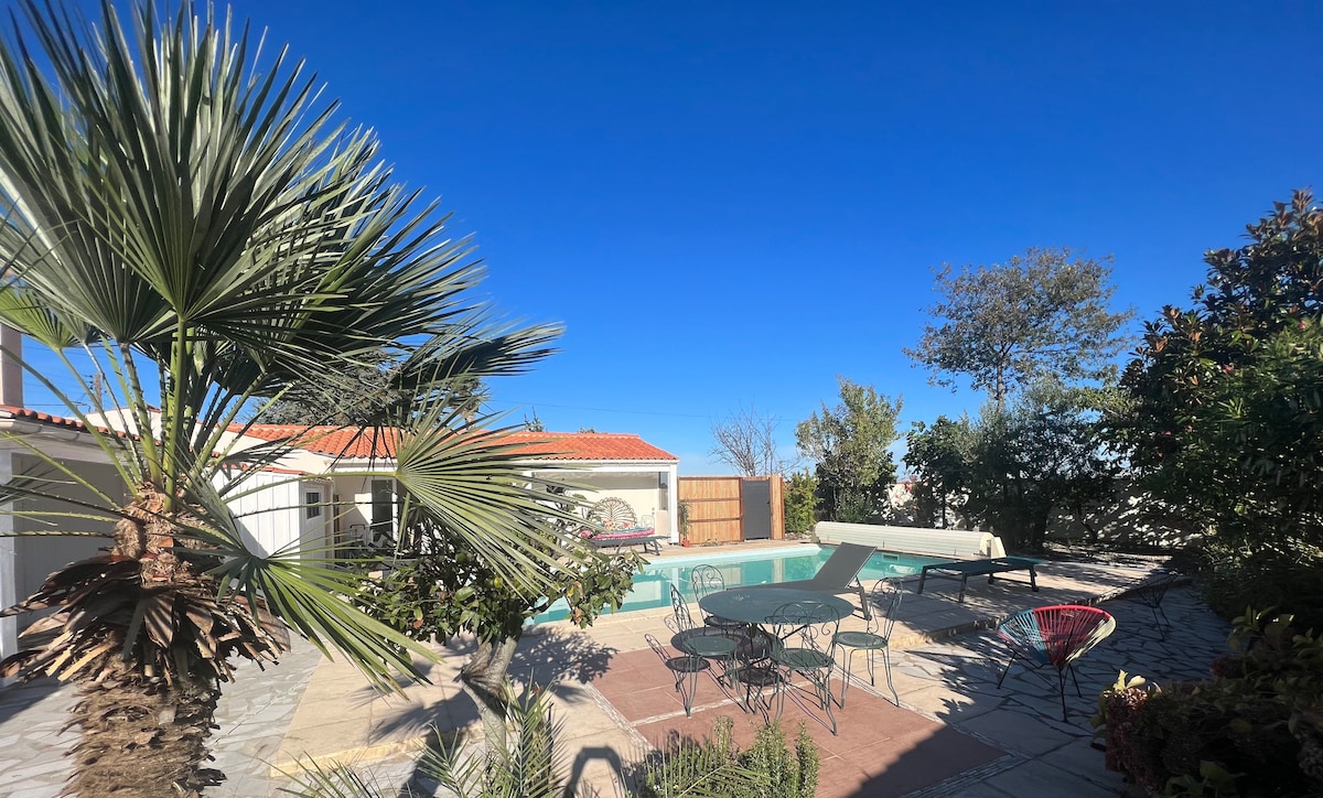A shared outdoor space features a rectangular pool surrounded by a natural stone patio. Several tables and chairs are arranged, accompanied by greenery including palm trees, offering a relaxing atmosphere under a clear blue sky.