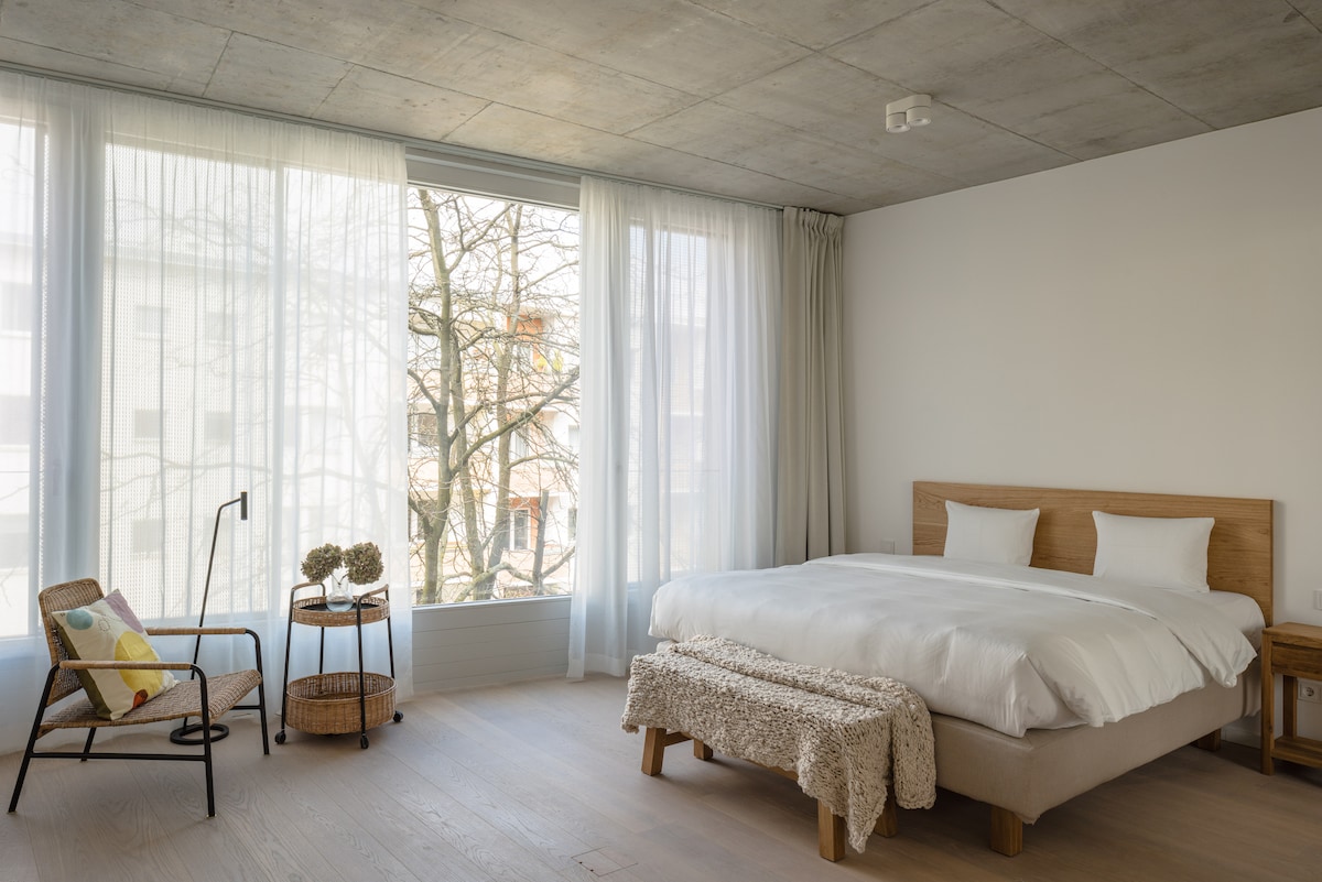 A spacious bedroom features a large bed with white linens, complemented by a textured blanket at the foot. A seating area includes a wooden chair next to a round table. Floor-to-ceiling windows allow natural light to enhance the neutral color scheme.