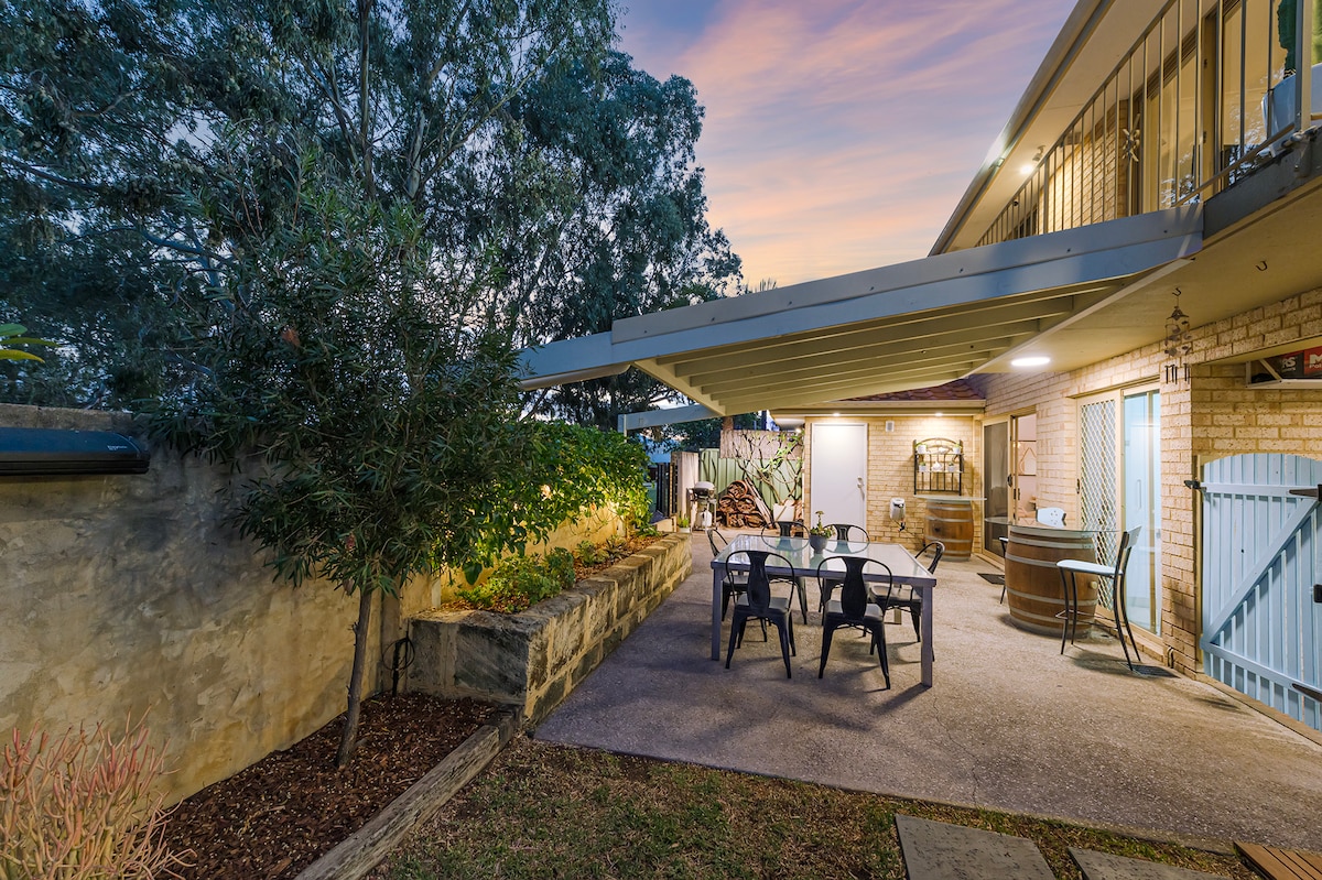 An outdoor patio area is visible, featuring a dining set with chairs and a table. A hot tub is positioned nearby, surrounded by greenery. The space is under a covered awning, with warm evening light illuminating the setting, creating a serene atmosphere for relaxation.
