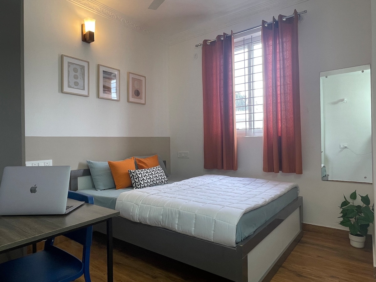 A bright and cozy room features a spacious bed with a light-colored comforter. Cushions in orange and a patterned black-and-white design are placed for comfort. A study table with a laptop is situated nearby, while natural light filters through a window with red curtains.