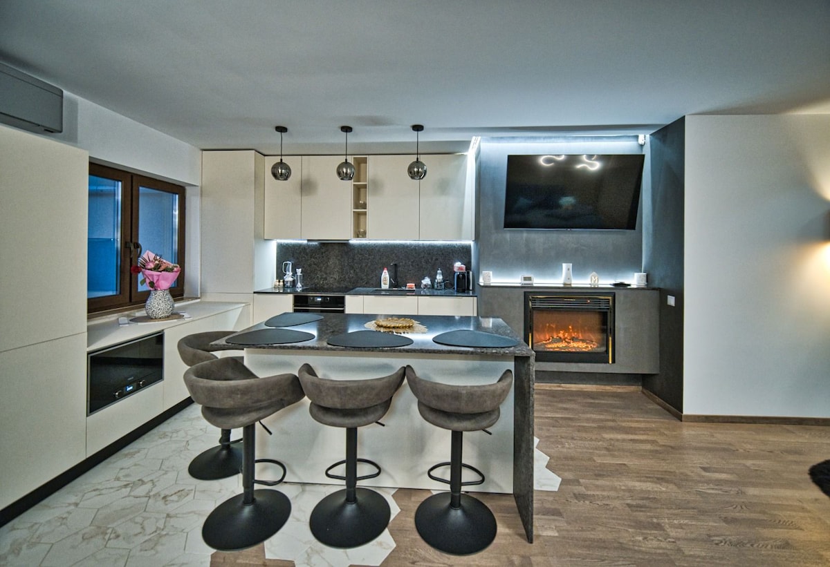 The kitchen area is equipped with modern appliances and features a large island with four high-backed stools. A sleek countertop complements the cabinetry, while a flat-screen TV is mounted above a contemporary fireplace, creating a stylish yet functional space.