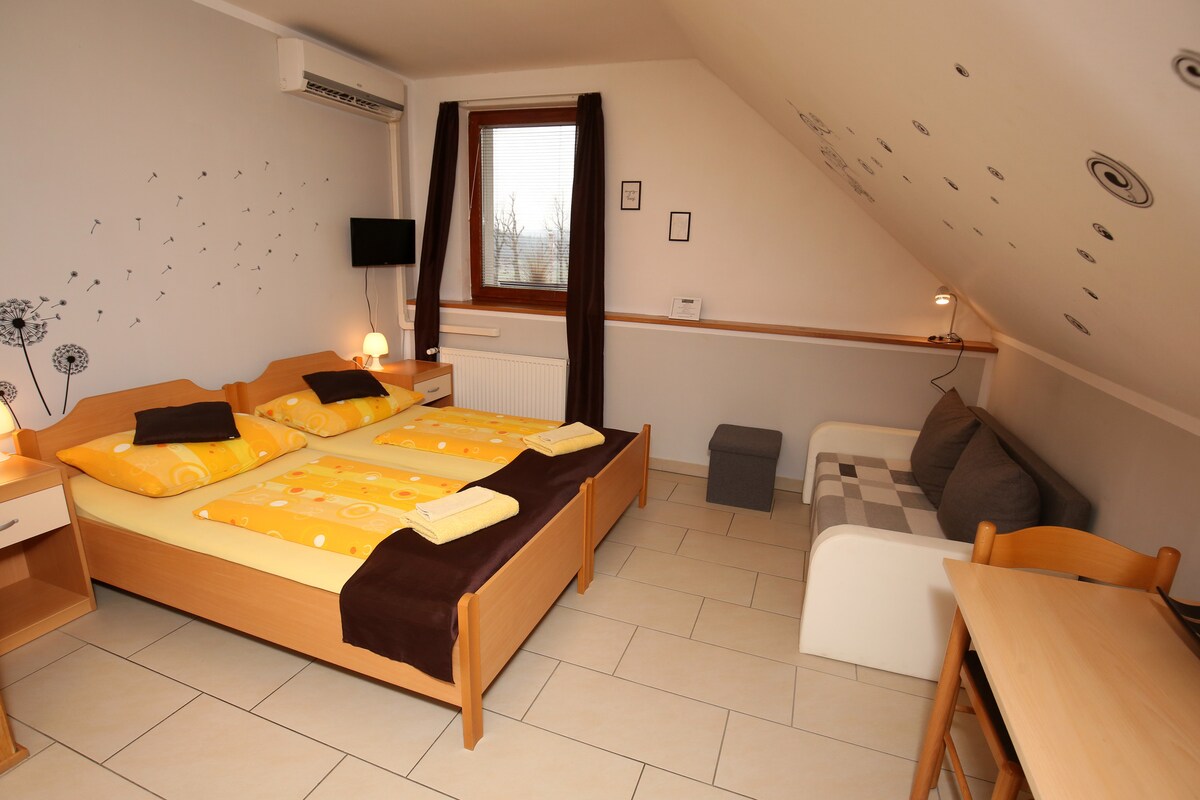 A bright and airy attic room features two spacious beds with colorful yellow bedding. A compact seating area includes a gray couch and a small ottoman. A desk with a wooden chair and a wall-mounted TV are also present. Natural light comes through a large window.