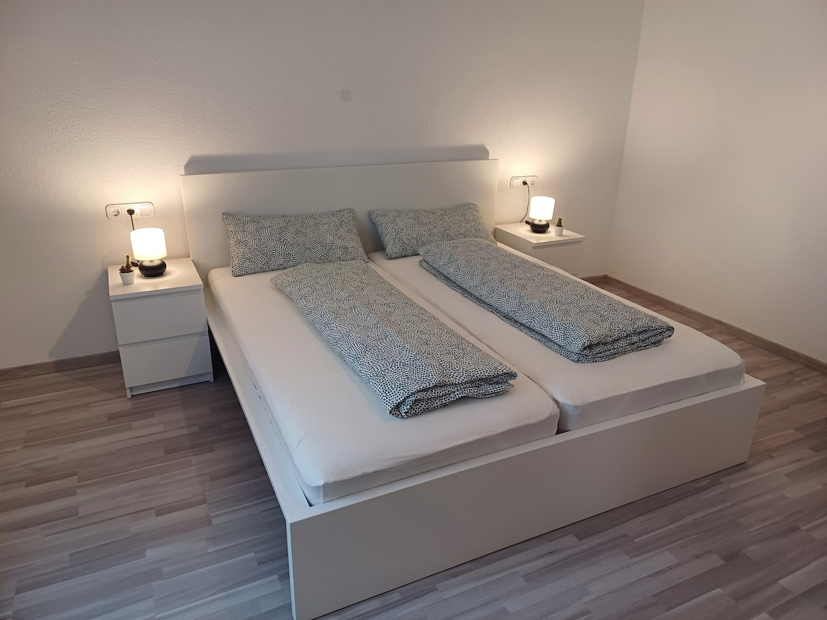 A minimalist bedroom is presented with a spacious bed featuring two separate mattresses. Each side table accommodates a lamp, providing balanced illumination. Light-colored walls and a soft wooden floor create a bright and airy setting, inviting relaxation.