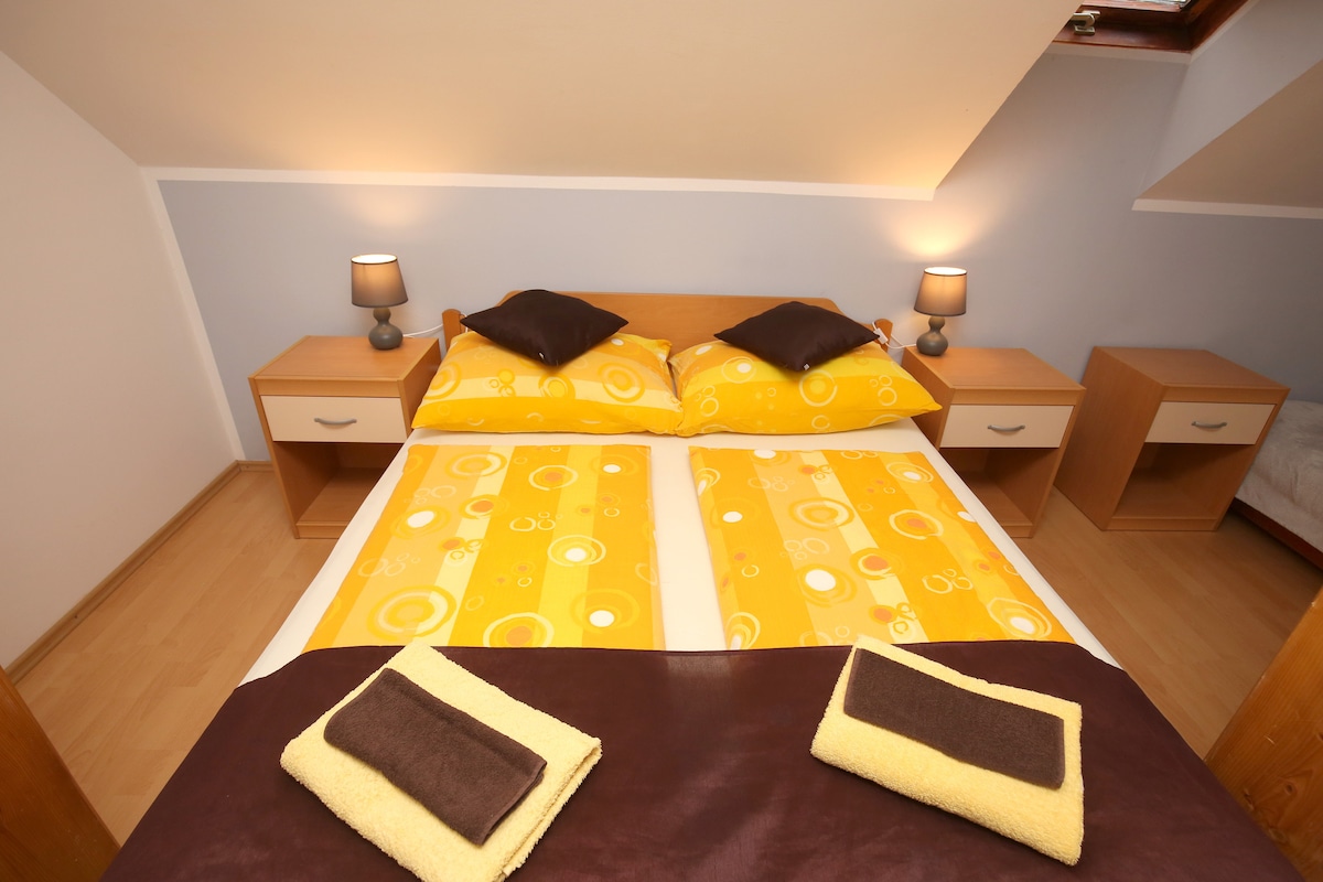 A well-lit sleeping area features a spacious bed with a bright yellow and white patterned duvet, complemented by two dark brown pillows. Nightstands are positioned on either side, each topped with lamps. Fresh towels are neatly arranged at the foot of the bed.