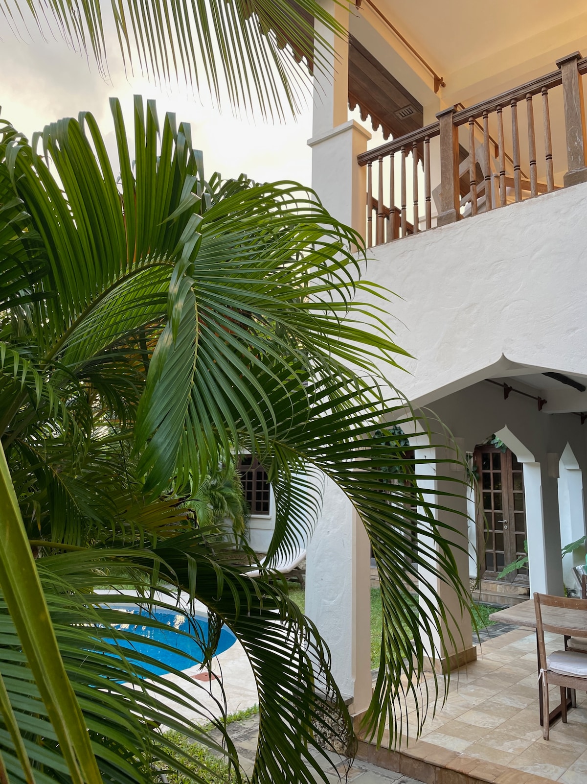 Lush palm leaves frame the view of the villa's exterior, showcasing a two-story structure with intricate architectural details. A serene swimming pool is visible in the background, surrounded by tile flooring and tropical greenery.