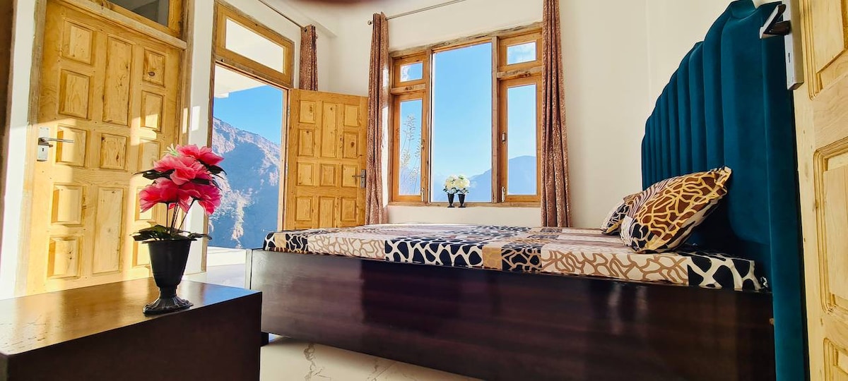 A bright bedroom features a spacious bed adorned with patterned bedding. Large windows frame stunning mountain views and allow natural light to fill the space. A bedside table holds a small vase with flowers, complementing the warm wooden accents throughout the room.