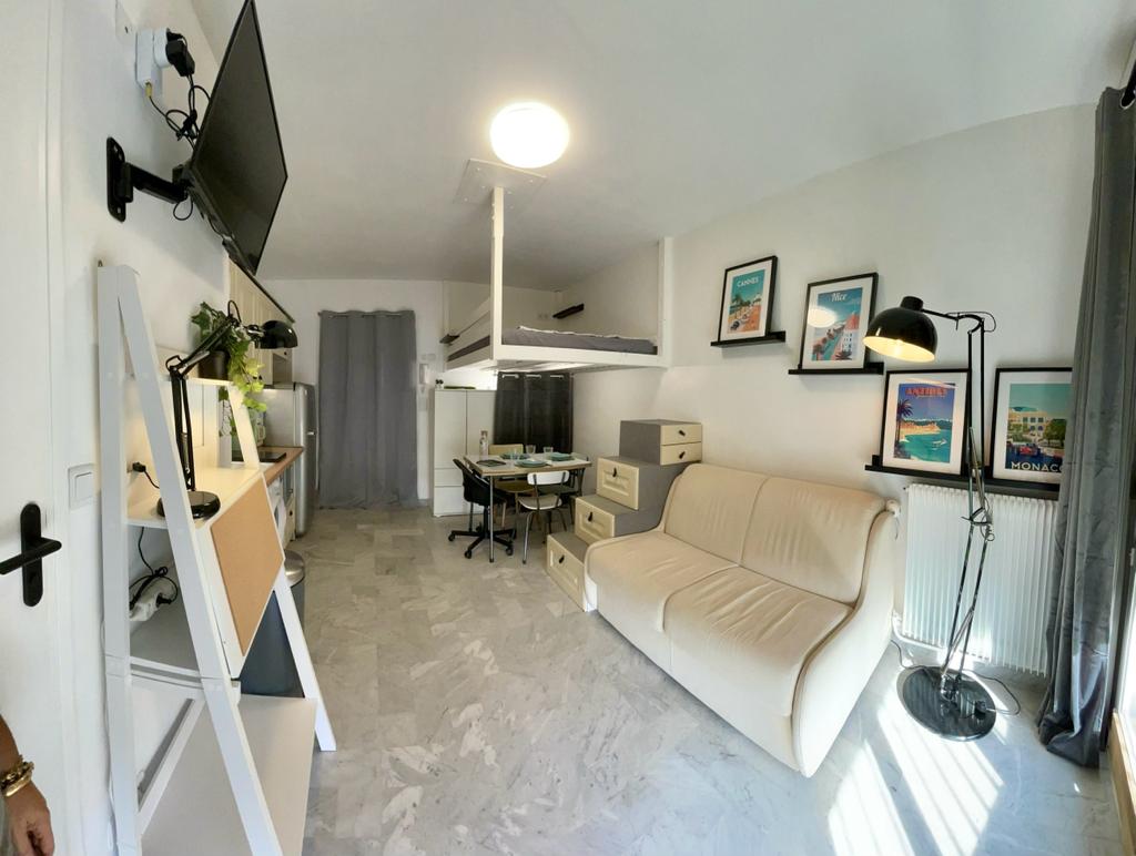 An open studio layout features a comfortable sofa and a work area with a table and chairs. Modern décor is complemented by artwork on the walls, and a light fixture illuminates the space. A kitchenette is visible, enhancing the functionality of the room.