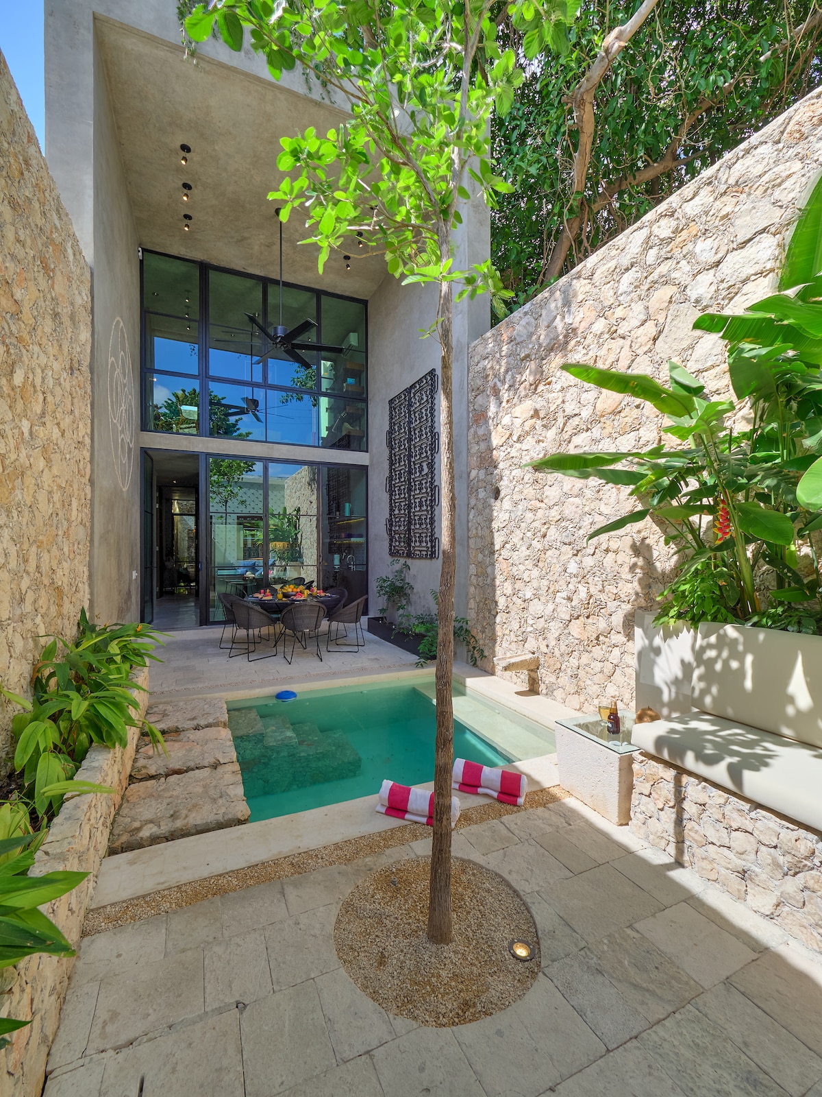 An open-air courtyard showcases a small pool surrounded by natural stone walls and vibrant greenery. A dining table sits nearby, inviting al fresco meals. Sunlight filters through a large glass wall, connecting the space with the interior of the house.