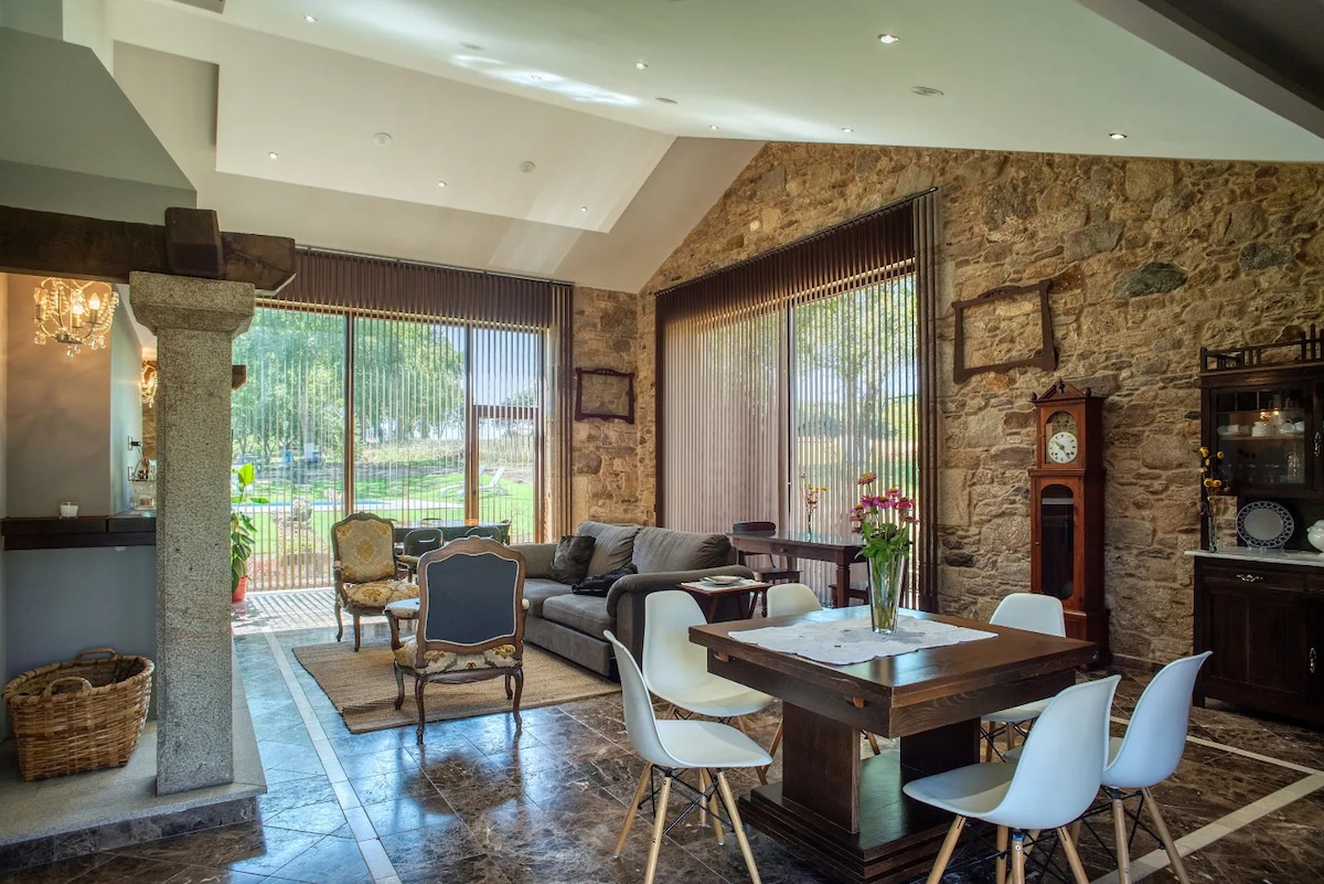 A spacious living area features large windows that allow natural light to fill the room. Exposed stone walls add texture, while a comfortable sofa and wooden dining table create a welcoming setting. A clock stands prominently, and potted plants are visible through the glass doors.