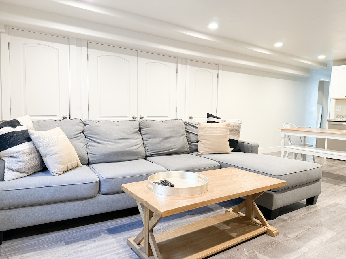 A comfortable sitting area is featured, highlighted by a spacious sectional couch adorned with decorative pillows. A wooden coffee table with a decorative tray sits in front, creating a relaxed space for gatherings. Light walls enhance the brightness of the room.