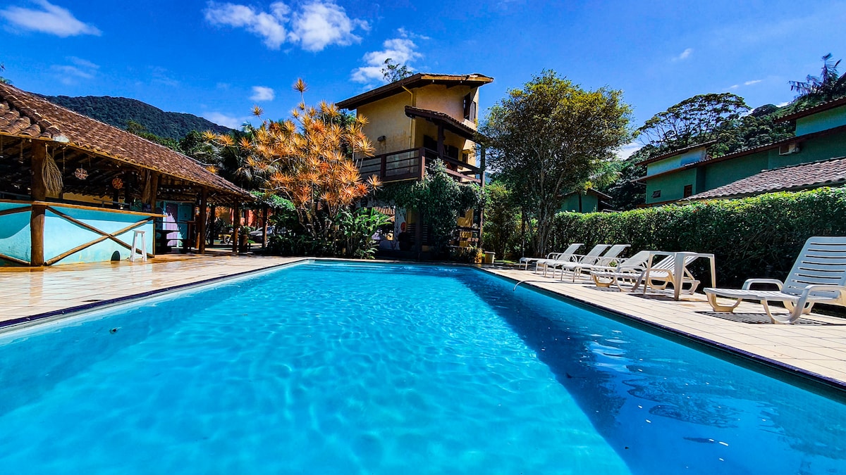 An outdoor pool is set within a spacious area framed by greenery and mountains. Lounge chairs are arranged around the pool deck, while a two-story building is visible in the background. Bright blue skies and scattered clouds enhance the serene atmosphere.