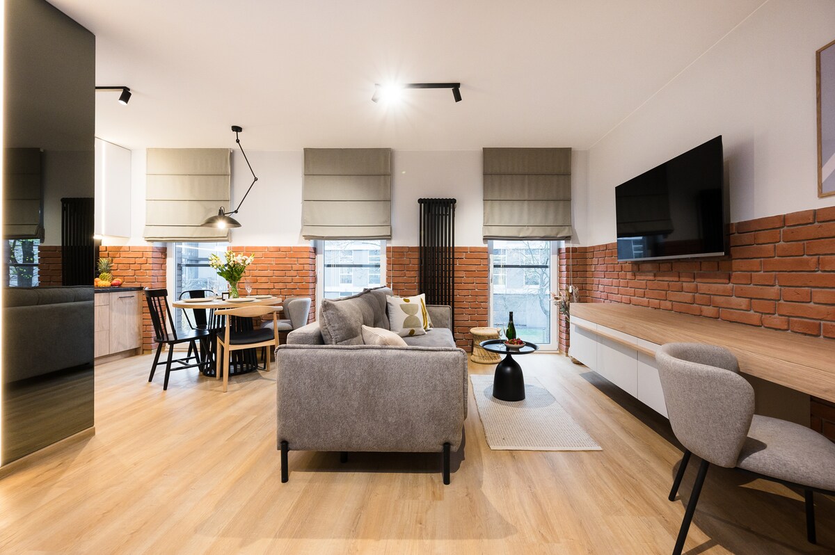 The living area features a modern design with a comfortable gray sofa and a round coffee table. Large windows with elegant blinds provide natural light. A dining table with black chairs is positioned nearby against a backdrop of exposed brick walls, enhancing the room's character.