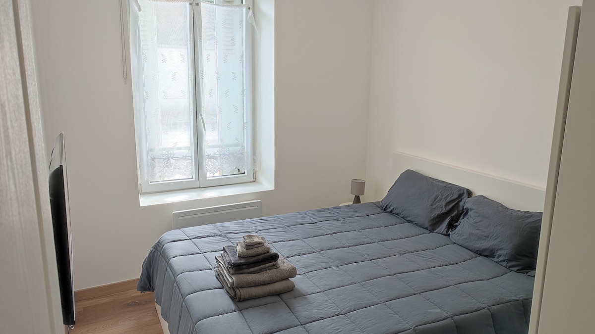 A simple and bright bedroom features a spacious bed with a textured blue coverlet. Stacked neatly on the bed are several gray towels. Natural light enters through a window with sheer curtains, creating a calm ambiance in the room.