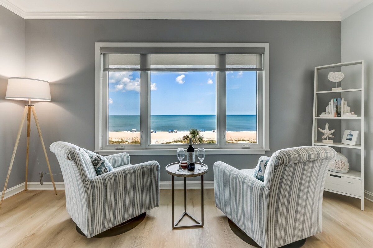 A cozy seating area is positioned beside a large window, offering an unobstructed view of the ocean. Two striped armchairs face a small round table. Natural light fills the space, highlighting the soft grey walls and light-colored wood flooring.