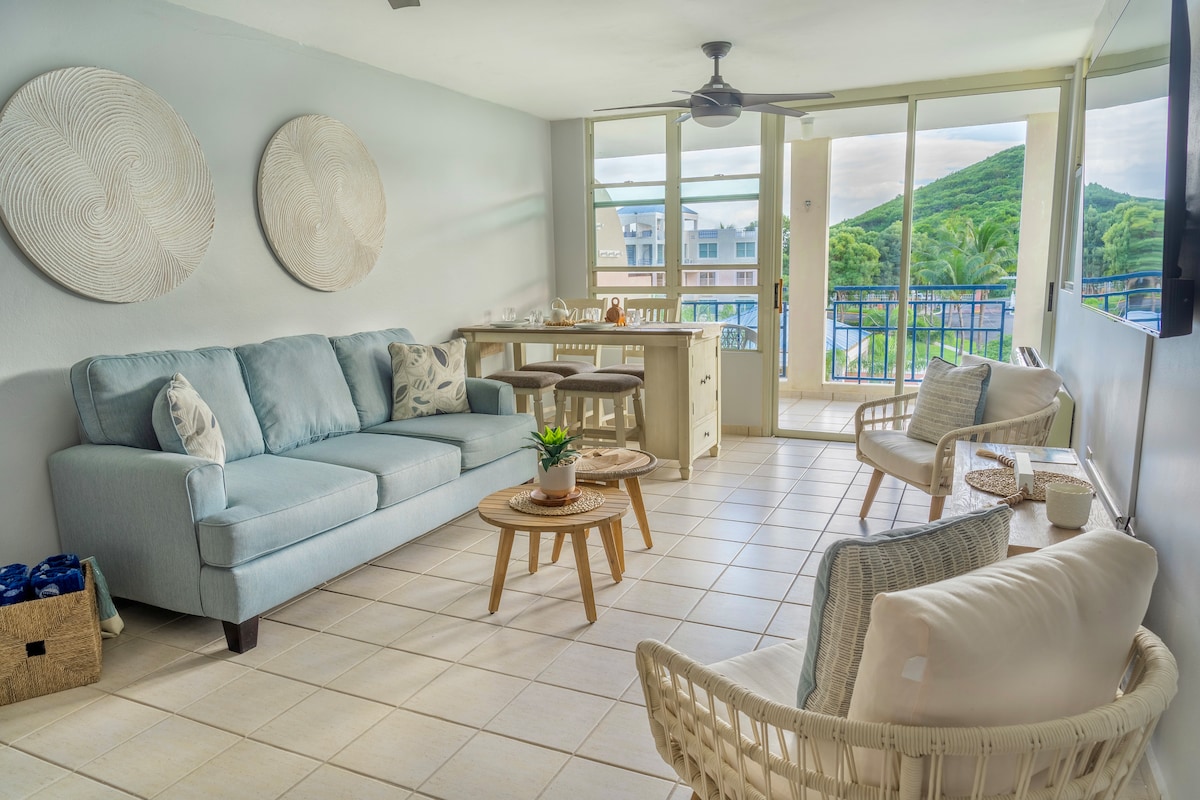 A comfortable living area features a light blue sofa, two woven chairs, and two small wooden tables. Large windows provide views of the surrounding greenery and allow ample natural light to fill the space. Neutral tones create a calm atmosphere, enhanced by decorative wall art.
