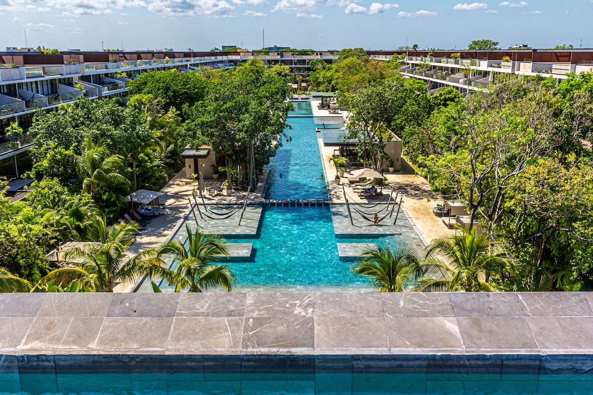 A spacious swimming pool area is surrounded by lush greenery, featuring multiple interconnected pools that extend through the landscape. Loungers are positioned throughout for relaxation, with palm trees adding a tropical touch to the serene environment.